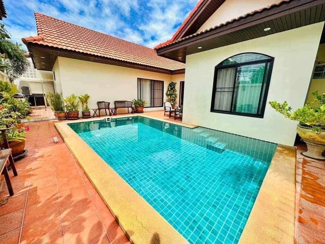 A serene private swimming pool is framed by lush greenery and terracotta tiles. The inviting water reflects the clear blue sky, while comfortable seating areas with wooden chairs are visible along the poolside. The villa's exterior features large windows and a tiled roof.