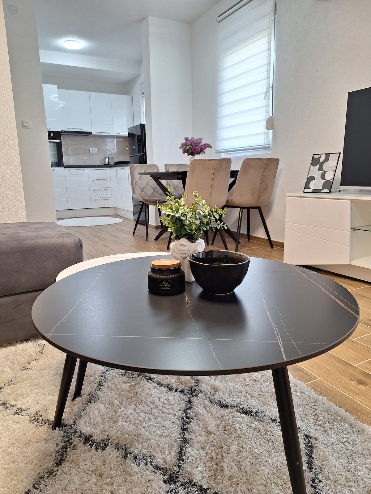 A modern black coffee table is positioned on a patterned rug, displaying a small decorative bowl and a candle. In the background, a dining area is visible with a set of beige chairs around a table, complemented by a well-equipped kitchen featuring white cabinetry.