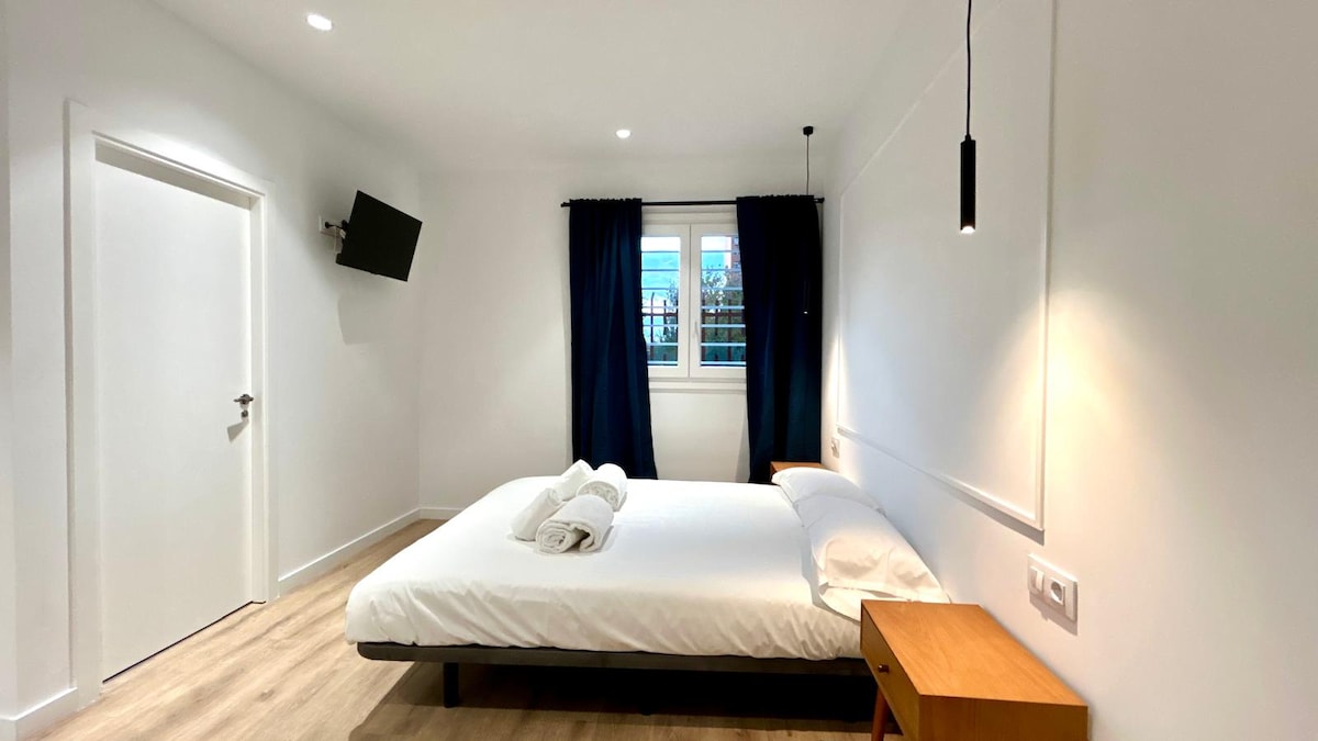 A bright studio room features a spacious bed with crisp linens, positioned against a neutral wall. A wall-mounted TV is visible, along with a stylish bedside table. Natural light enters through a window dressed in dark curtains, creating a calm and relaxing environment.