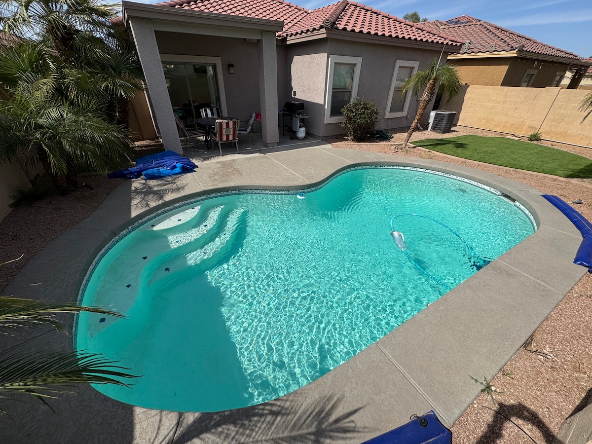 A sparkling pool occupies the outdoor area, featuring a unique shape and clear turquoise water. Surrounding the pool, a concrete deck is complemented by palm plants and a grassy lawn. The patio area, visible in the background, includes seating options and a gas grill.
