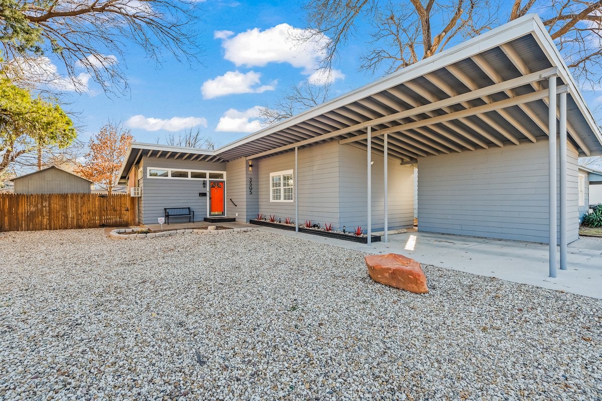 The exterior features a modern single-story home with a light gray facade and a welcoming bright orange front door. A spacious gravel area is surrounded by low-maintenance landscaping and a covered carport. The property is set against a backdrop of trees and a wooden fence.