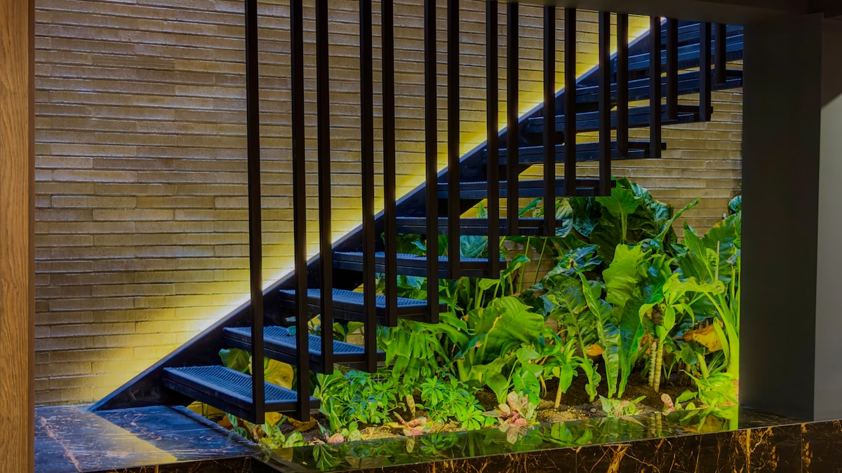 A modern staircase features sleek black metal steps, gracefully illuminated by soft lighting. Surrounding the base, a variety of lush green plants creates a vibrant contrast against the textured stone wall, enhancing the contemporary aesthetic of the space.