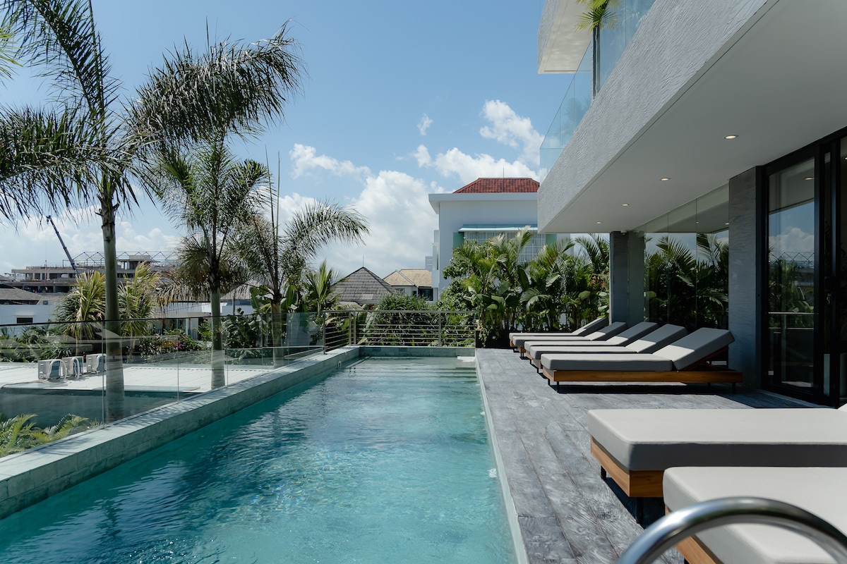 A modern pool area is highlighted by a sleek infinity pool surrounded by lush tropical landscaping. Sun loungers are positioned along the edge, offering a relaxing spot to enjoy the serene environment under a clear blue sky.