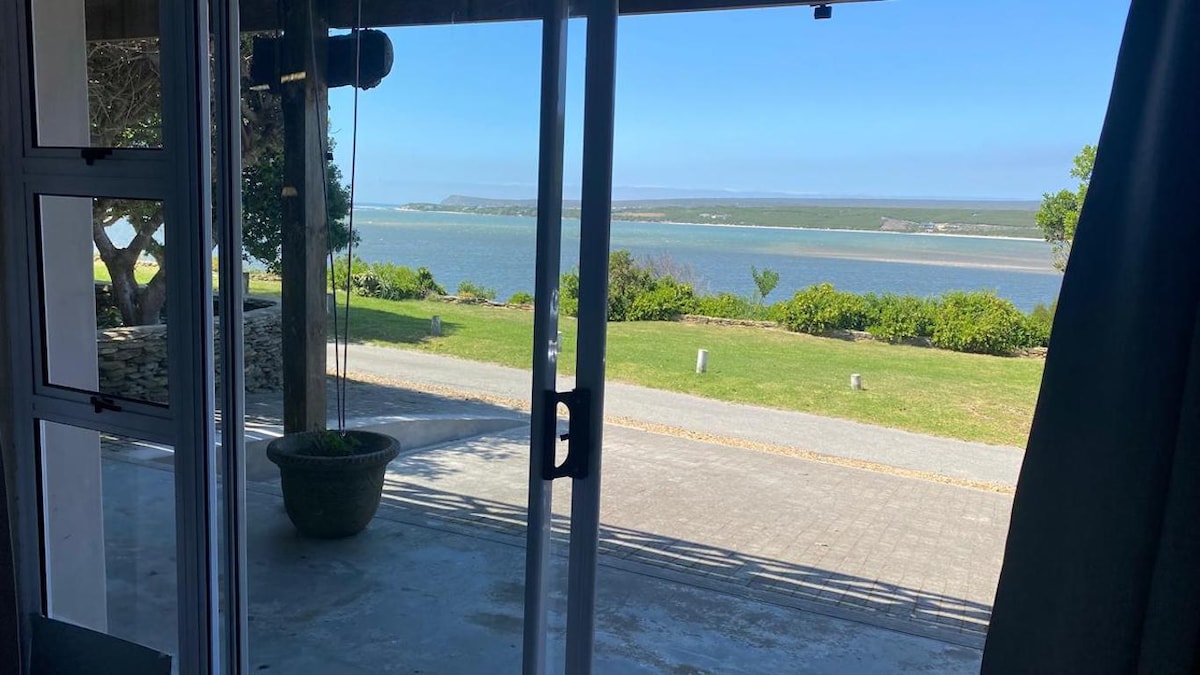 A view of the tranquil Breede River is framed by large glass doors, inviting natural light into the space. Lush greenery and the expansive river are visible in the distance, enhancing the serene outdoor atmosphere.