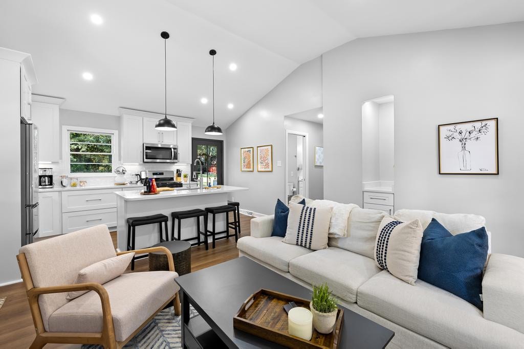 A modern living area features a light sofa adorned with decorative pillows, accompanied by a wooden armchair. A dark coffee table sits in the center, displaying a small plant. The open kitchen is visible in the background, equipped with stainless steel appliances and a spacious island.