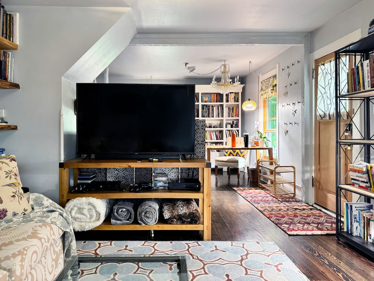 A welcoming living area features a large television on a wooden media stand. A bookshelf filled with books is situated against a wall, and a cozy work space is positioned near a window. Stylish textiles adorn the floor, adding warmth to the space.