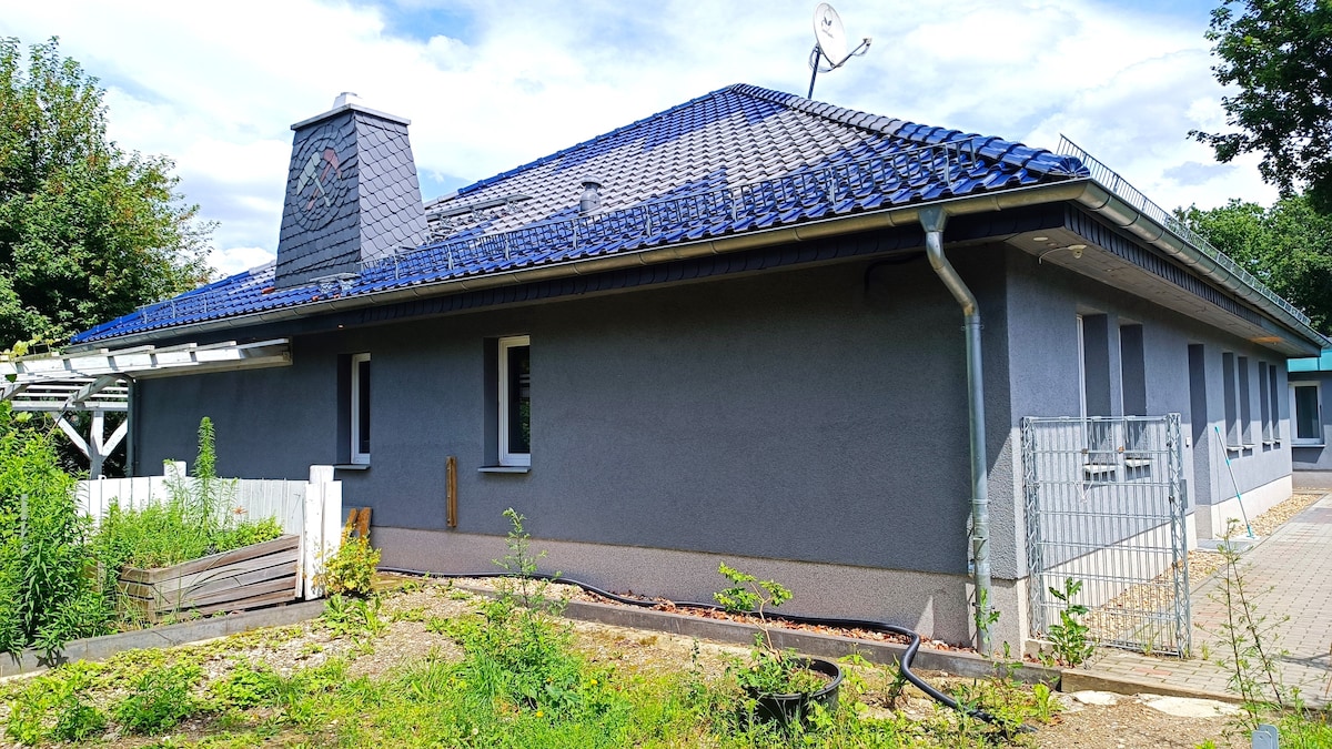 The exterior of the property features a modern structure with a blue tiled roof and grey walls. Windows are spaced along the side, and a fenced area with greenery is visible in the foreground, providing a calm and natural feel to the setting.