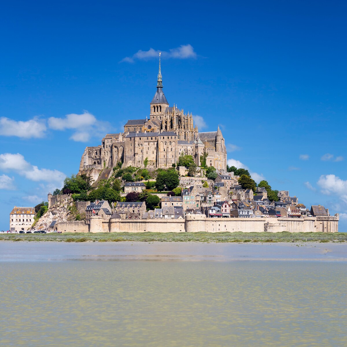 A historical island commune rises majestically from the water, featuring a towering abbey at its peak. Its medieval architecture is complemented by sloping hills and coastal vegetation, while quaint buildings line the base, creating a striking contrast against the bright blue sky.