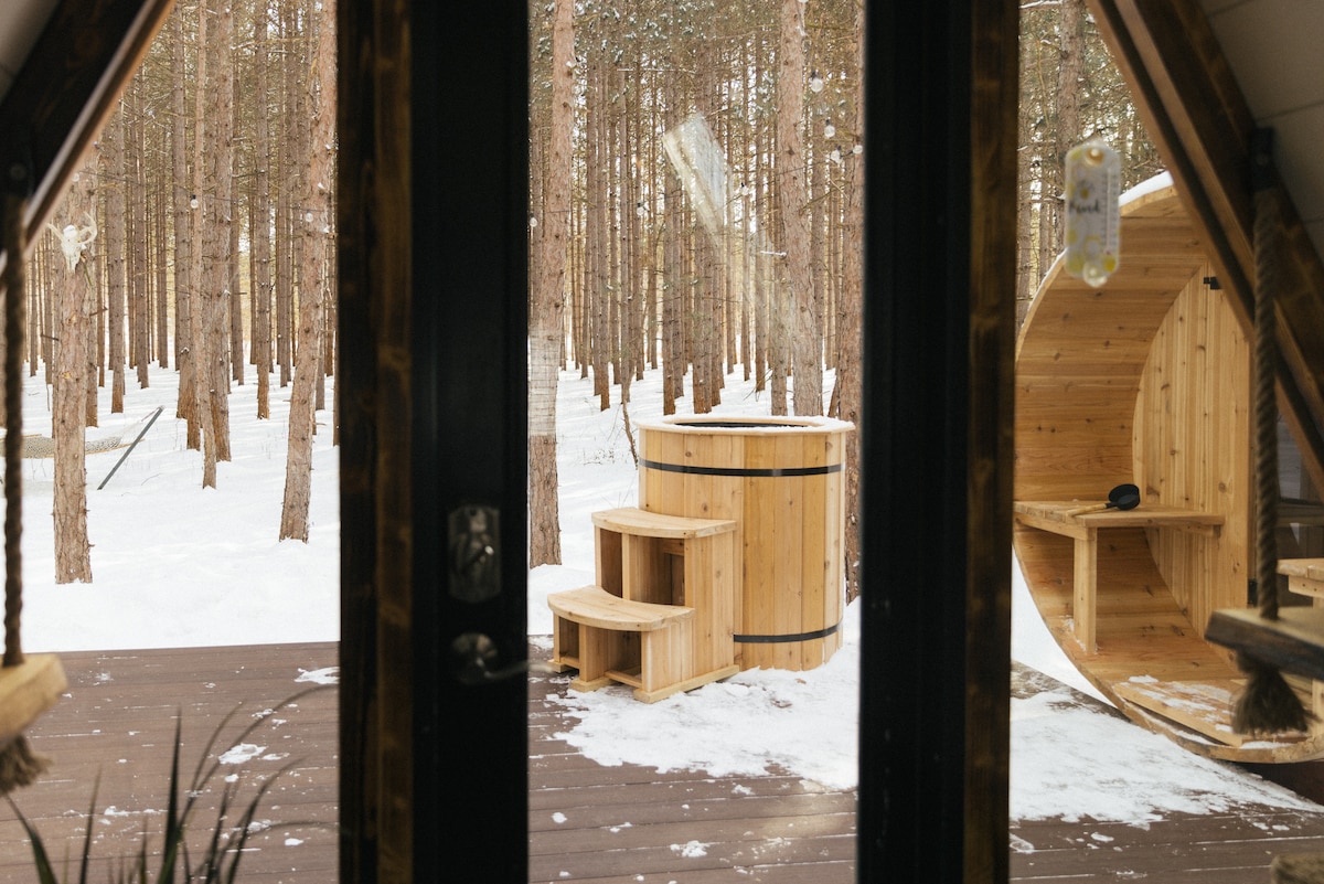 A view from the entrance showcases the outdoor deck, featuring a cedar barrel sauna and a cold plunge tub surrounded by trees blanketed in snow. Natural wood textures complement the tranquil winter setting, providing a secluded atmosphere.