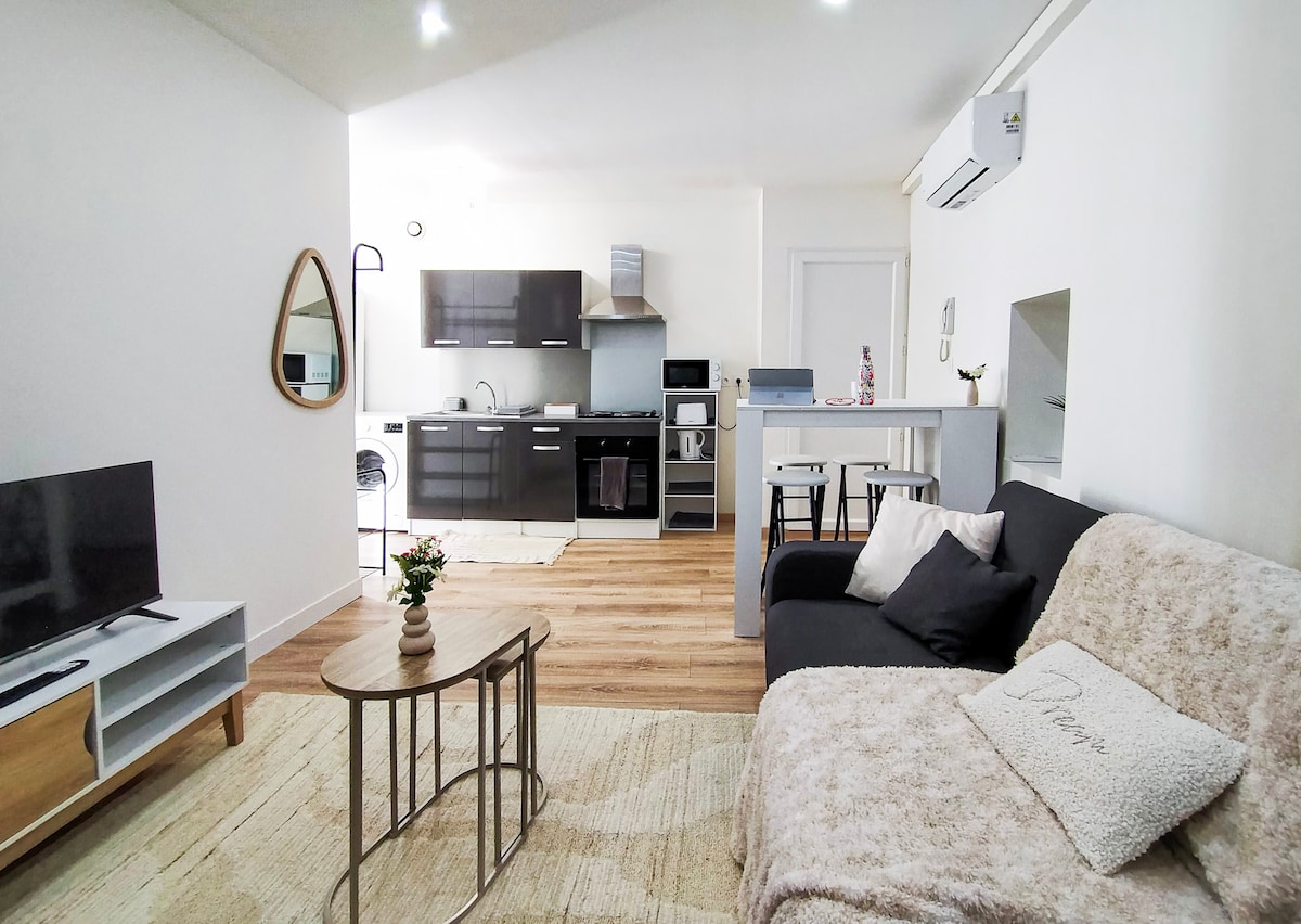 An open living area features a modern kitchen with sleek cabinetry and essential appliances, complemented by a comfortable sofa in front of a flat-screen television. A round coffee table and dining setup provide functionality, while natural light fills the space through the windows.