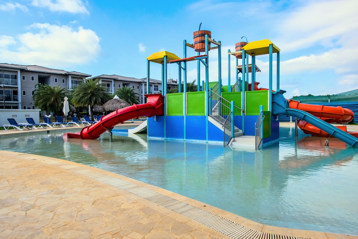 A colorful water play structure stands prominently in the shallow end of a pool, featuring two bright slides and interactive elements. Sun loungers are arranged neatly along the pool's edge, set against a backdrop of palm trees and a multi-story building.