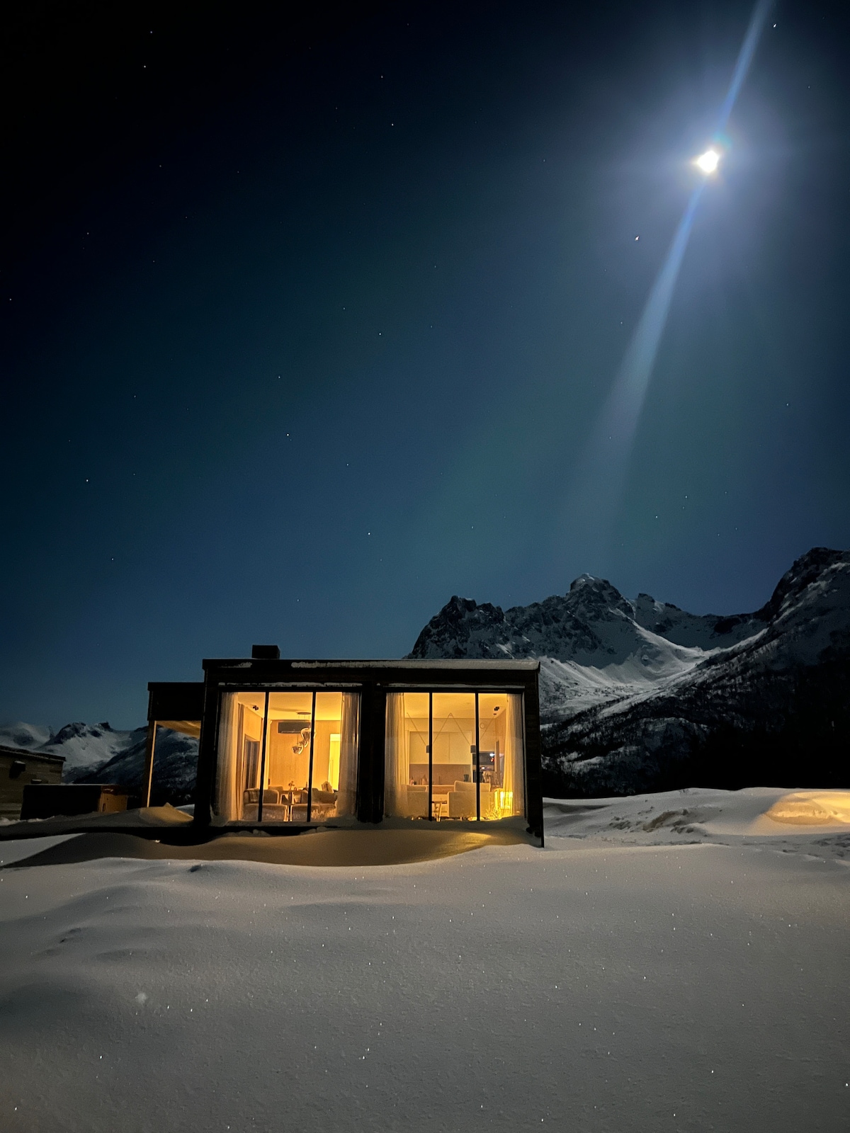 The modern cabin is illuminated from within, set against a night sky filled with stars. Snow blankets the ground, and mountains rise majestically in the background. A beam of moonlight highlights the structure, creating a serene atmosphere.