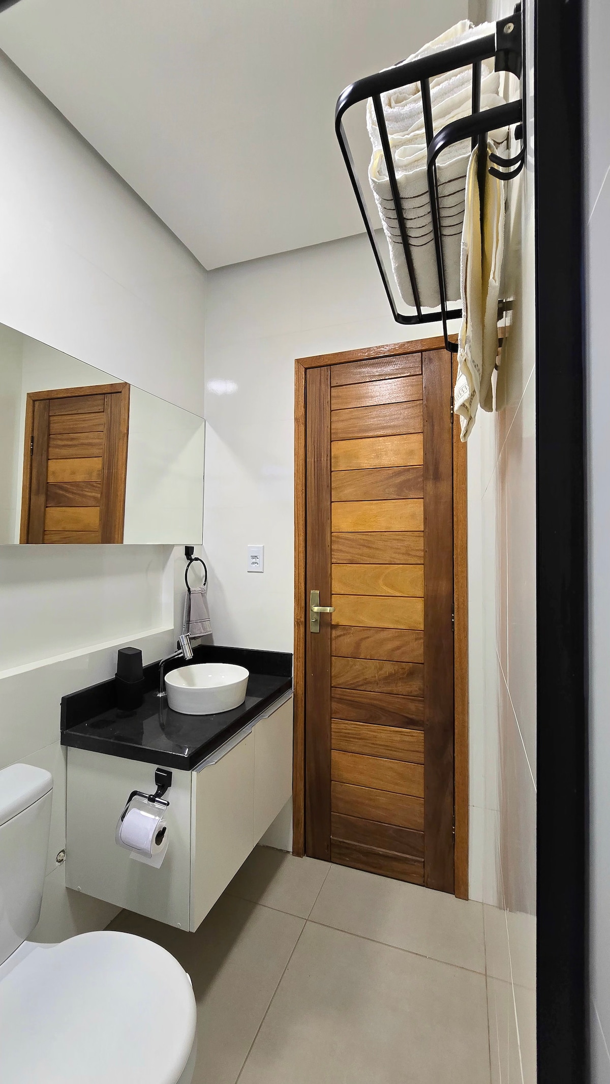 A modern bathroom is showcased, featuring a sleek black countertop with a round sink and a wall-mounted mirror. Wooden accents are seen on the door and towel rack, while organized towels are neatly arranged above the sink. A toilet is positioned adjacent to the counter.