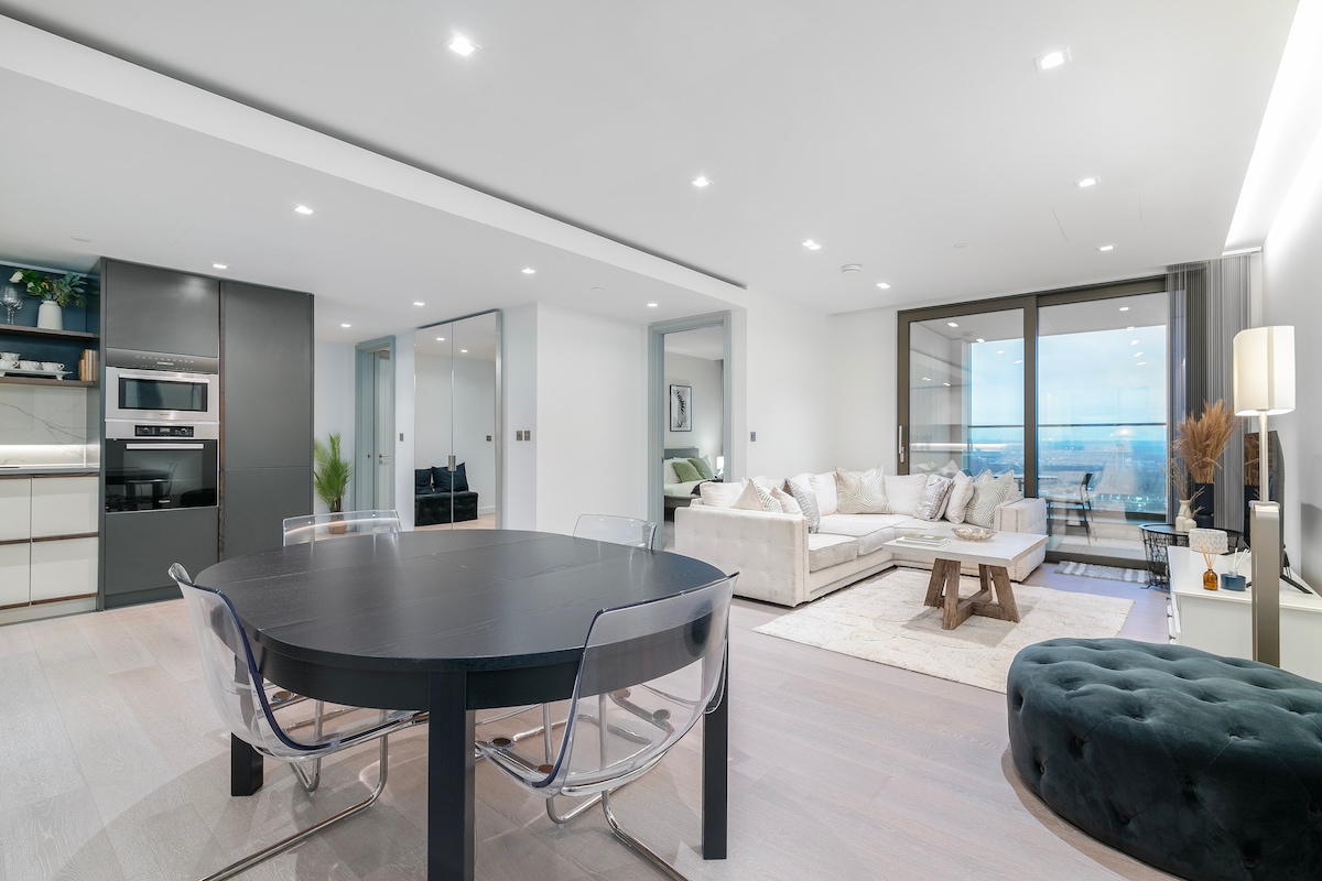 An open-plan living and dining area is illuminated by natural light from floor-to-ceiling windows, showcasing panoramic views. A round black dining table is surrounded by transparent chairs, while a spacious sofa, accented by cushions, offers a comfortable seating area. A plush ottoman is positioned nearby.