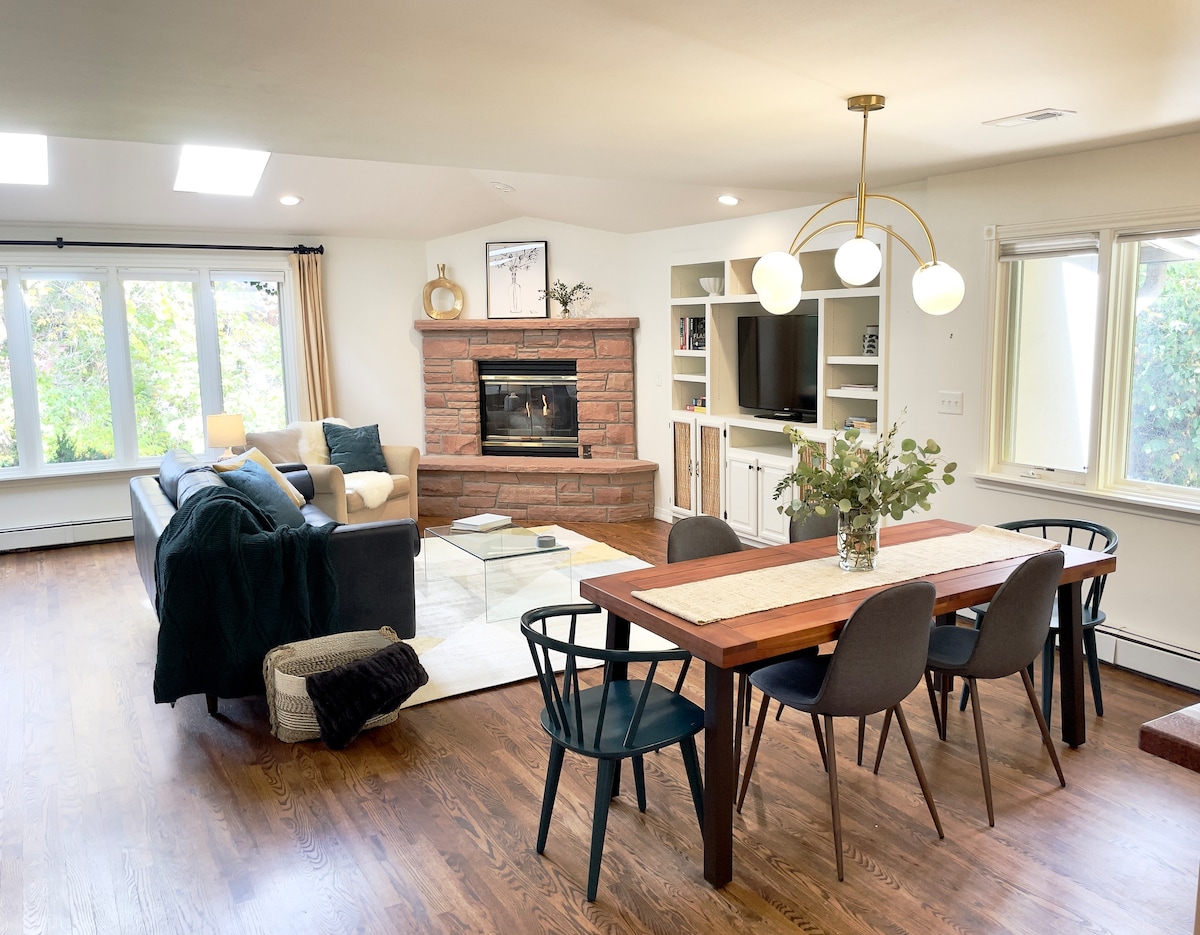 An open-plan living and dining area features a cozy gas fireplace set in a stone surround. A wooden dining table with black chairs is positioned near a window that offers views of greenery. A comfortable couch and decorative throw blanket enhance the space.