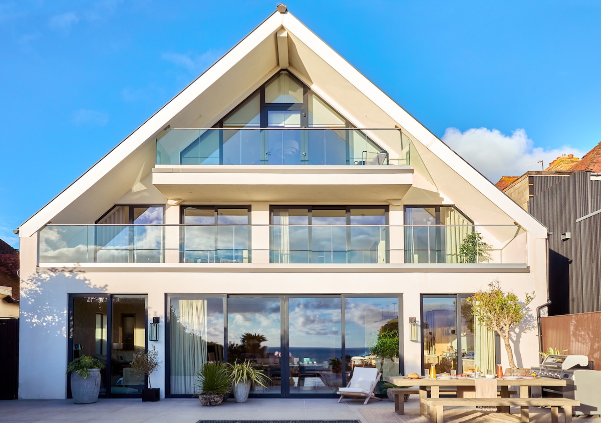 The contemporary exterior features a sloped roof and large glass balcony railings. Expansive windows allow natural light to illuminate the structure, while a stone patio with a wooden dining table and lush greenery adds a welcoming touch. Sea views are visible beyond the house.
