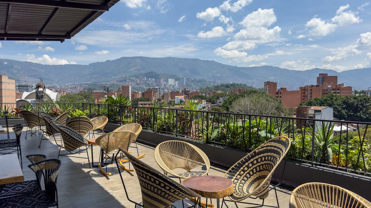 A spacious rooftop terrace is presented, featuring several rattan chairs arranged for seating. Lush greenery surrounds the area, with panoramic views of the mountains and cityscape visible in the background under a clear blue sky. Natural light enhances the inviting outdoor setting.
