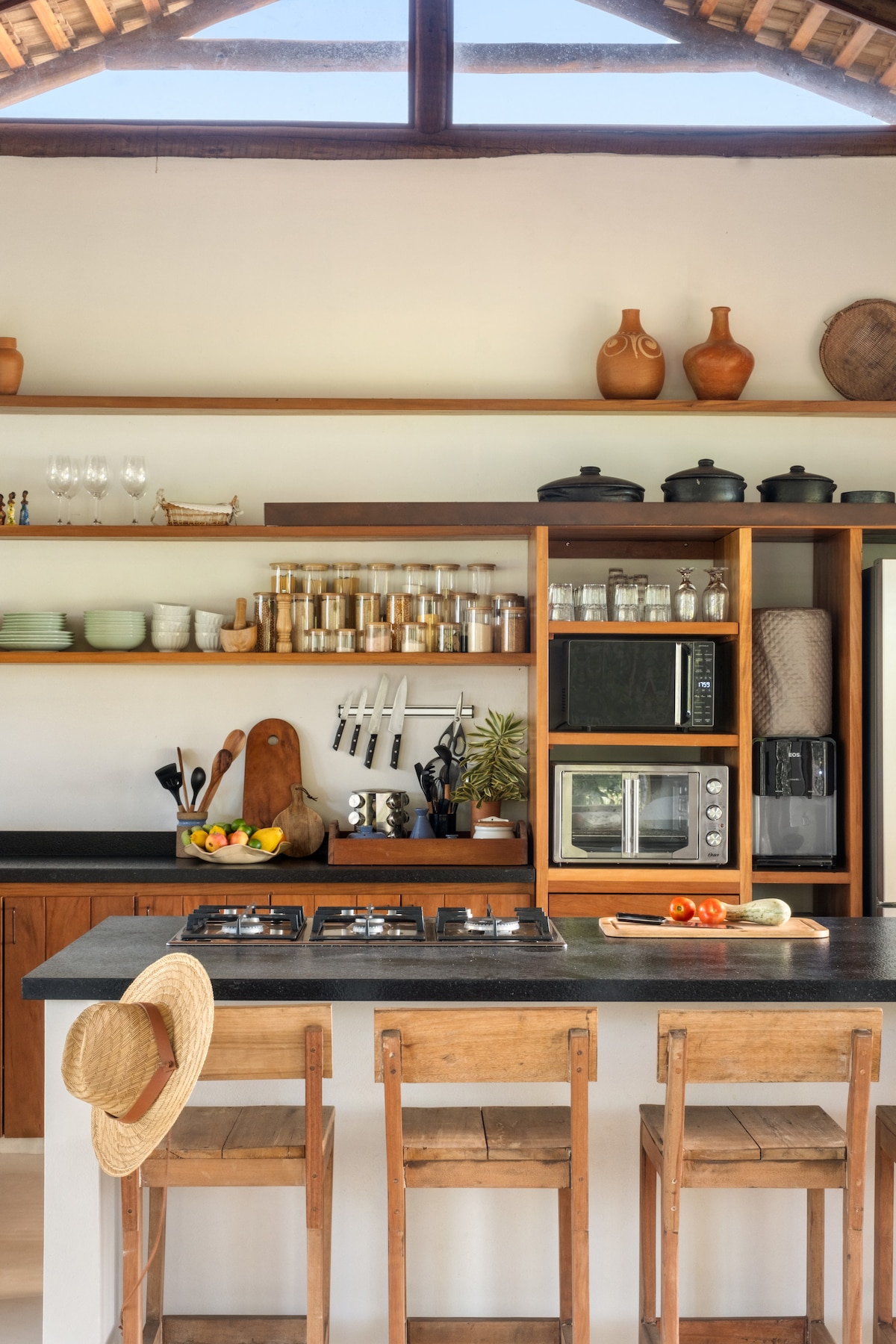 A modern kitchen features wooden shelves lined with glassware and ceramic dishes, complemented by a selection of pots and pans. A cooking area with a stovetop and a microwave is present, alongside wooden bar stools at a counter. Bright items, including fruits, are arranged for a fresh touch.