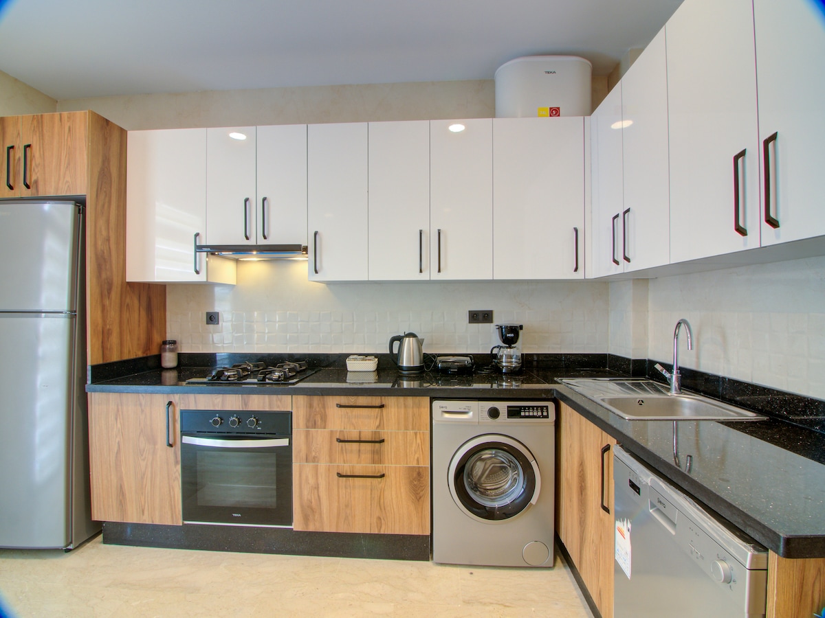 The kitchen features modern appliances, including a stainless steel refrigerator, oven, and washing machine. Dark countertops contrast with light cabinetry, creating a clean and functional workspace. A coffee maker and electric kettle are also present, enhancing the kitchen's usability for all cooking needs.