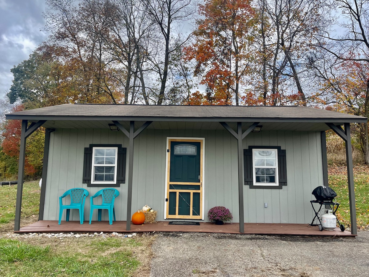 The exterior of a cozy cottage is shown, featuring a welcoming porch with two blue chairs and a small grill. A pumpkin and a potted mum decorate the entrance. Trees in autumn colors provide a backdrop, with a cloudy sky overhead.