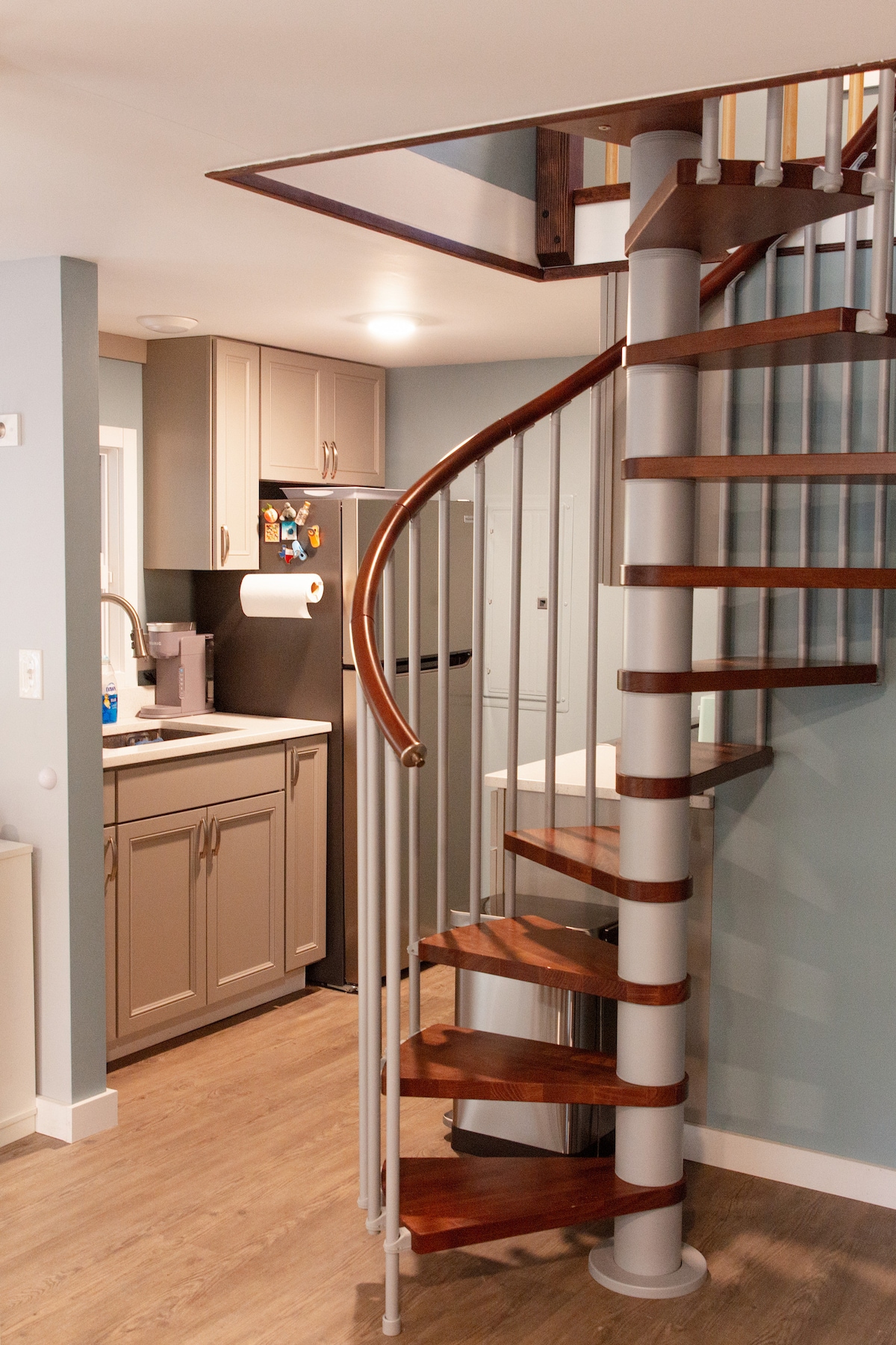 A modern spiral staircase, crafted from wood and metal, leads to the upper level of the cabin. The adjacent kitchen features light grey cabinets and a countertop, with a sink visible. Warm wood flooring adds a cozy touch to the environment.