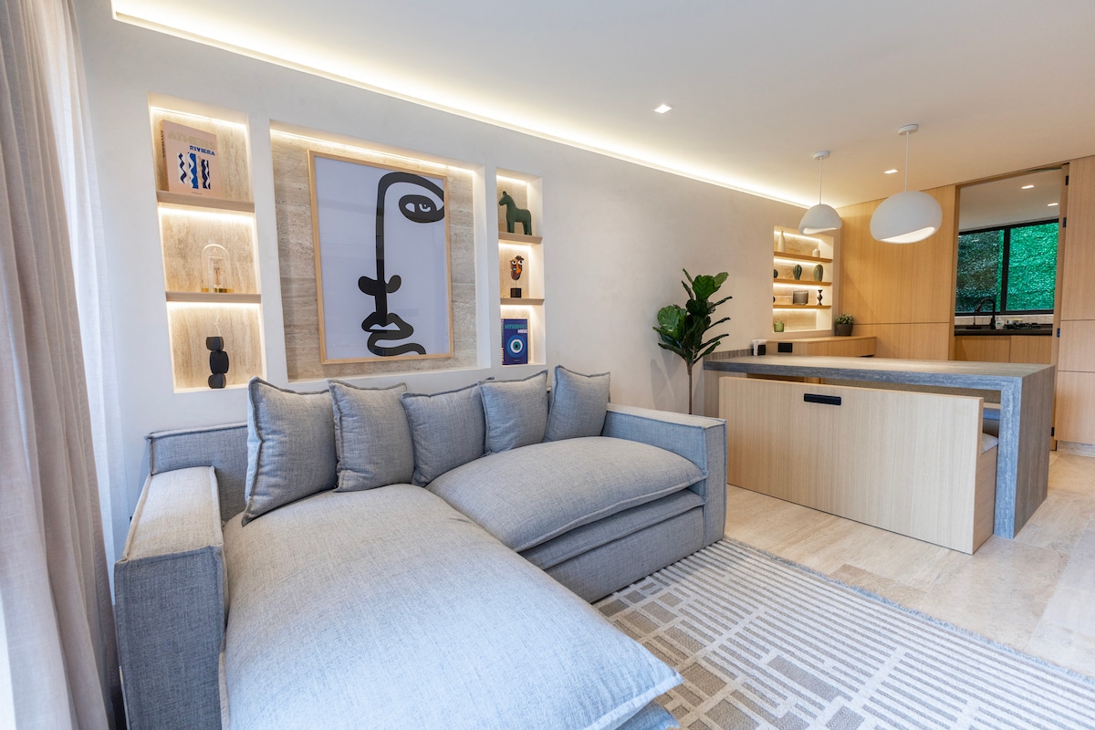 A modern living area is displayed, featuring a large sectional sofa with plush cushions. A light-hued rug lies beneath, complementing the soft tones of the walls. Decorative shelves are integrated into the side wall, showcasing various art pieces and plants. Natural light filters through nearby windows.