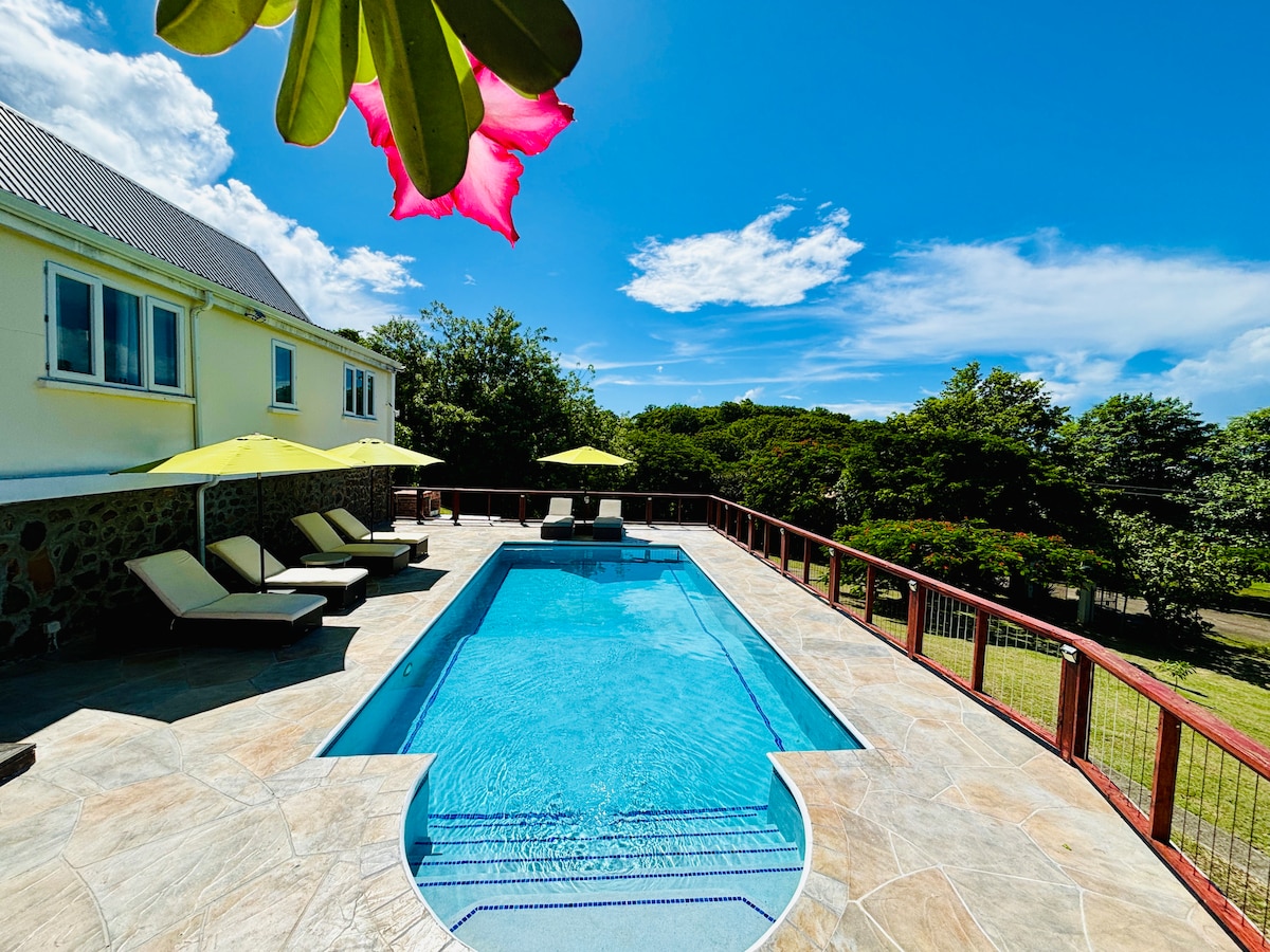 A serene outdoor space features a private swimming pool surrounded by a spacious deck. Lounge chairs with umbrellas are positioned for relaxation, while lush greenery outlines the area, creating a peaceful atmosphere under a clear blue sky.