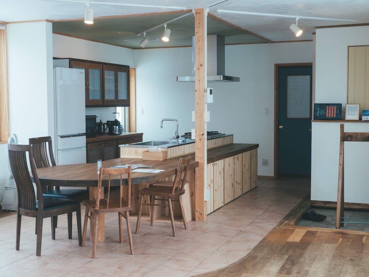 An open kitchen and dining area are visible, featuring a spacious island with a gas stove and various kitchen appliances, including a refrigerator and coffee maker. A wooden dining table is surrounded by four chairs. Natural light enters through windows, illuminating the tiled floor.