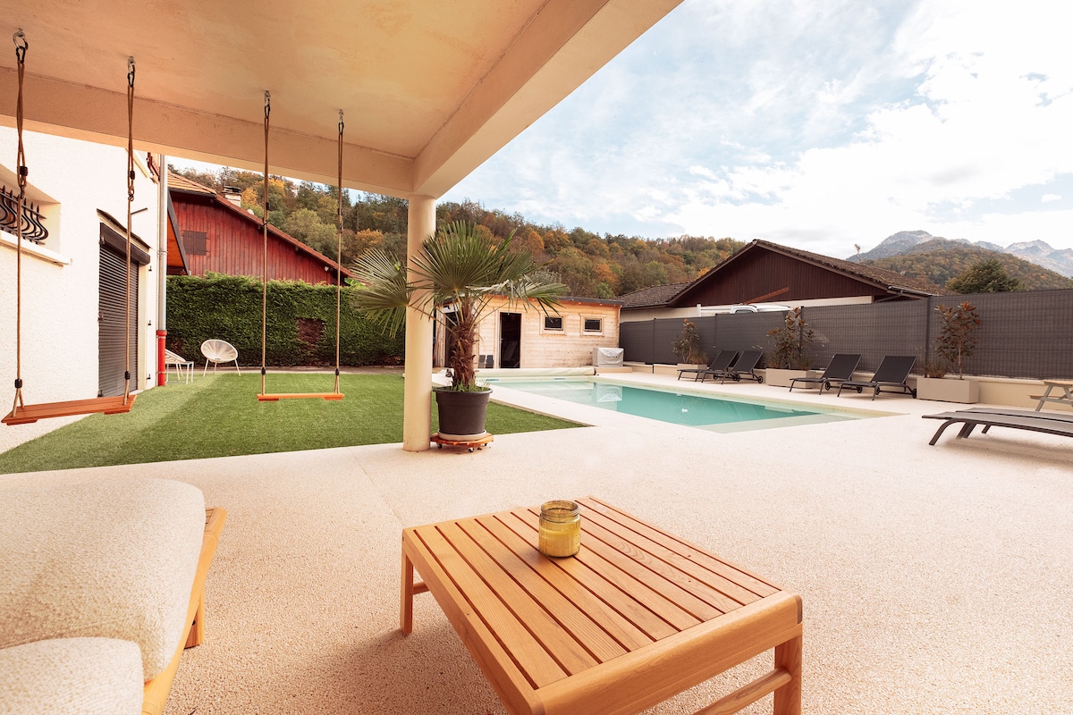 An outdoor area features a heated swimming pool surrounded by sun loungers, a wooden table, and a small seating area. Charming swings hang nearby, with lush greenery and distant mountains visible in the backdrop. A sense of tranquility is provided by the well-maintained garden.