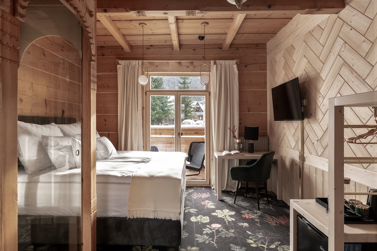 A wooden-paneled room features a spacious bed adorned with soft bedding. A balcony door leads to outdoor views. A sitting area includes a green chair and small table, while a television is mounted on the wall. Decorative floral carpet complements the rustic design.