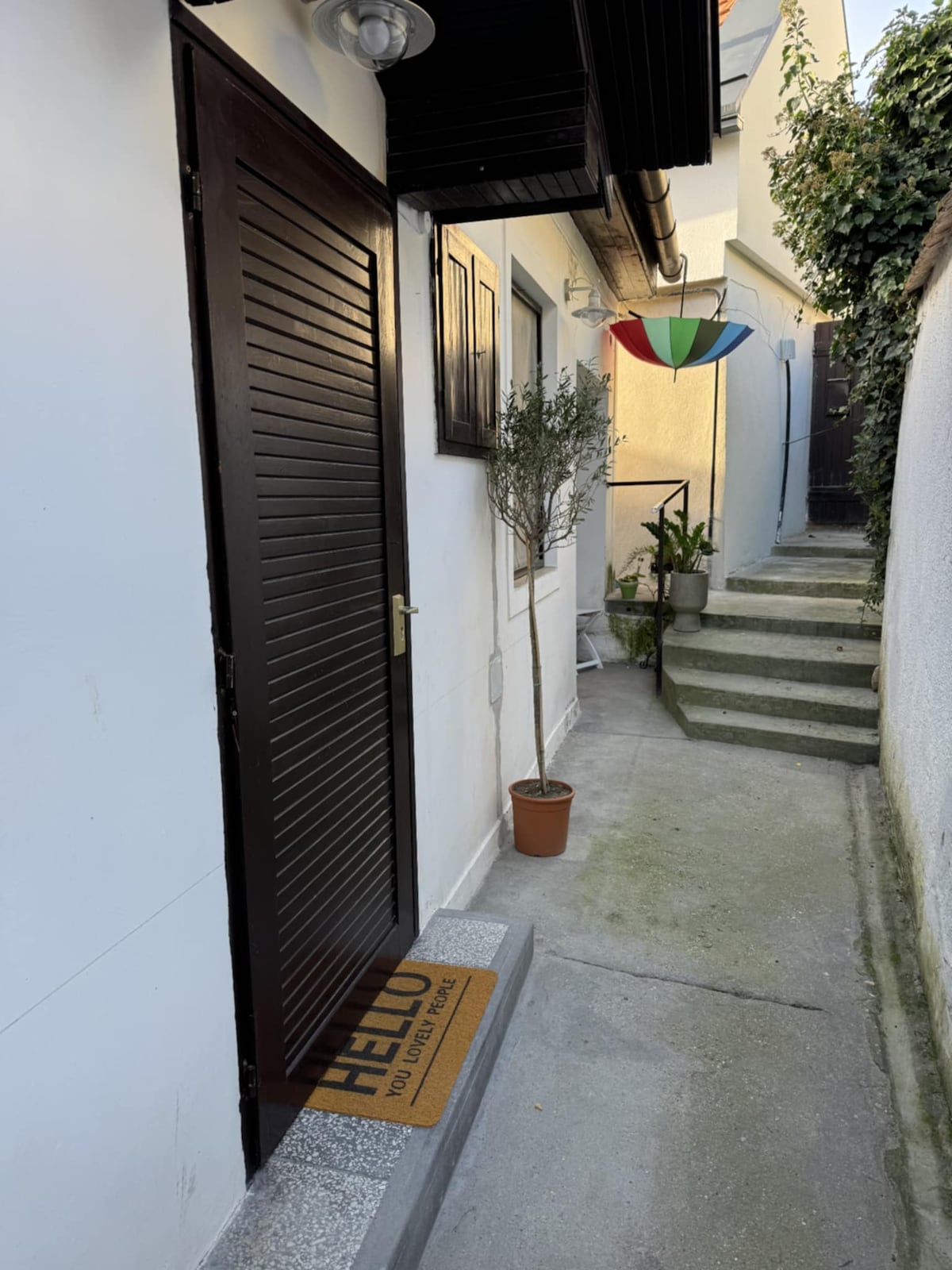 A narrow pathway leads to a wooden door, framed by a small potted olive tree and a vibrant umbrella overhead. The clean surroundings feature a stone step entrance, with steps visible leading toward the back. A welcome mat is placed at the door, inviting visitors.