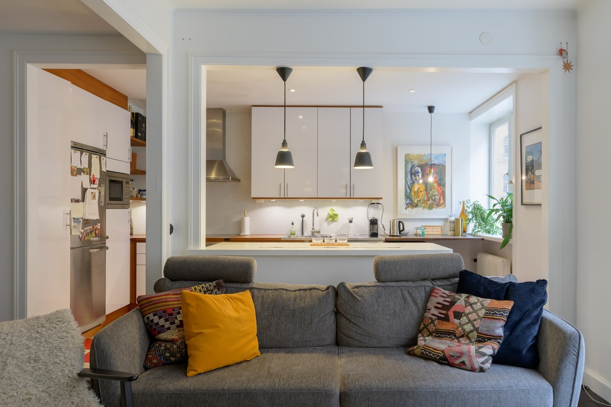 The living area features a dark gray sofa adorned with vibrant pillows, providing a cozy seating option. A well-equipped kitchen is visible in the background, showcasing modern appliances and clean, minimalist cabinetry. Two pendant lights hang above the kitchen space, adding a subtle warmth.
