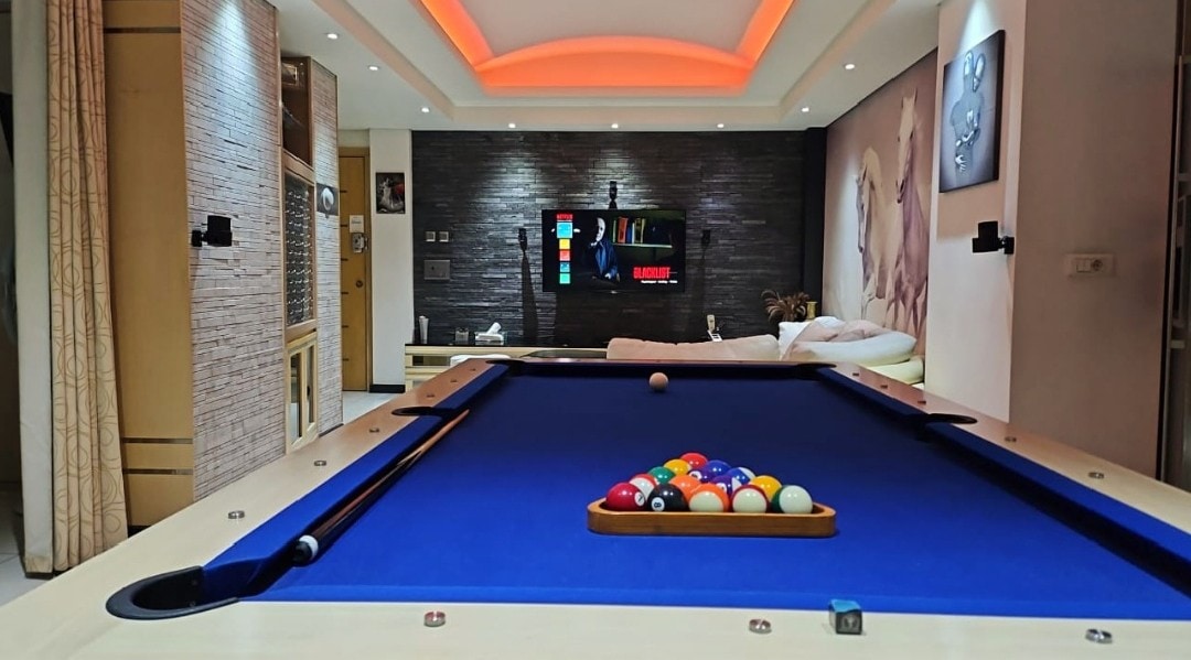 A professional pool table is situated in the dining area, featuring a blue felt surface and a triangle of colorful balls positioned for play. A sleek TV mount and modern decor in the background contribute to the inviting atmosphere.
