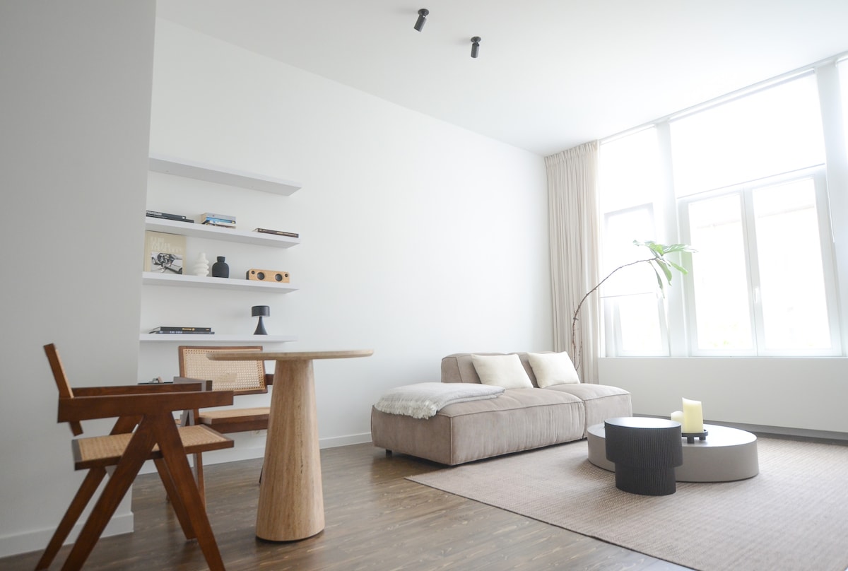 A light, airy living space features a modern sofa and a round black coffee table at its center. A wooden dining table with a design chair is positioned nearby. Natural light streams in through large windows, highlighting the minimalist decor and soft color palette.