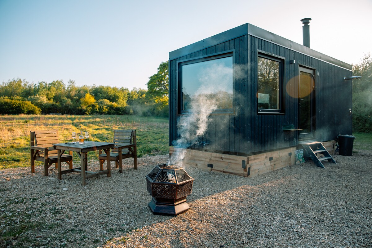 A modern off-grid cabin is shown amongst an open meadow. A picnic table sits nearby, accompanied by a fire pit with gentle smoke rising. Large windows reflect natural light, enhancing the connection with the surrounding landscape.