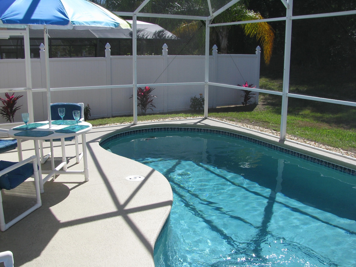 A private pool area is showcased within a screened enclosure, featuring clear blue water and a semi-circular design. Two white chairs and a small table are placed beside the pool, complemented by an umbrella. Lush greenery is visible in the background, enhancing the serene environment.