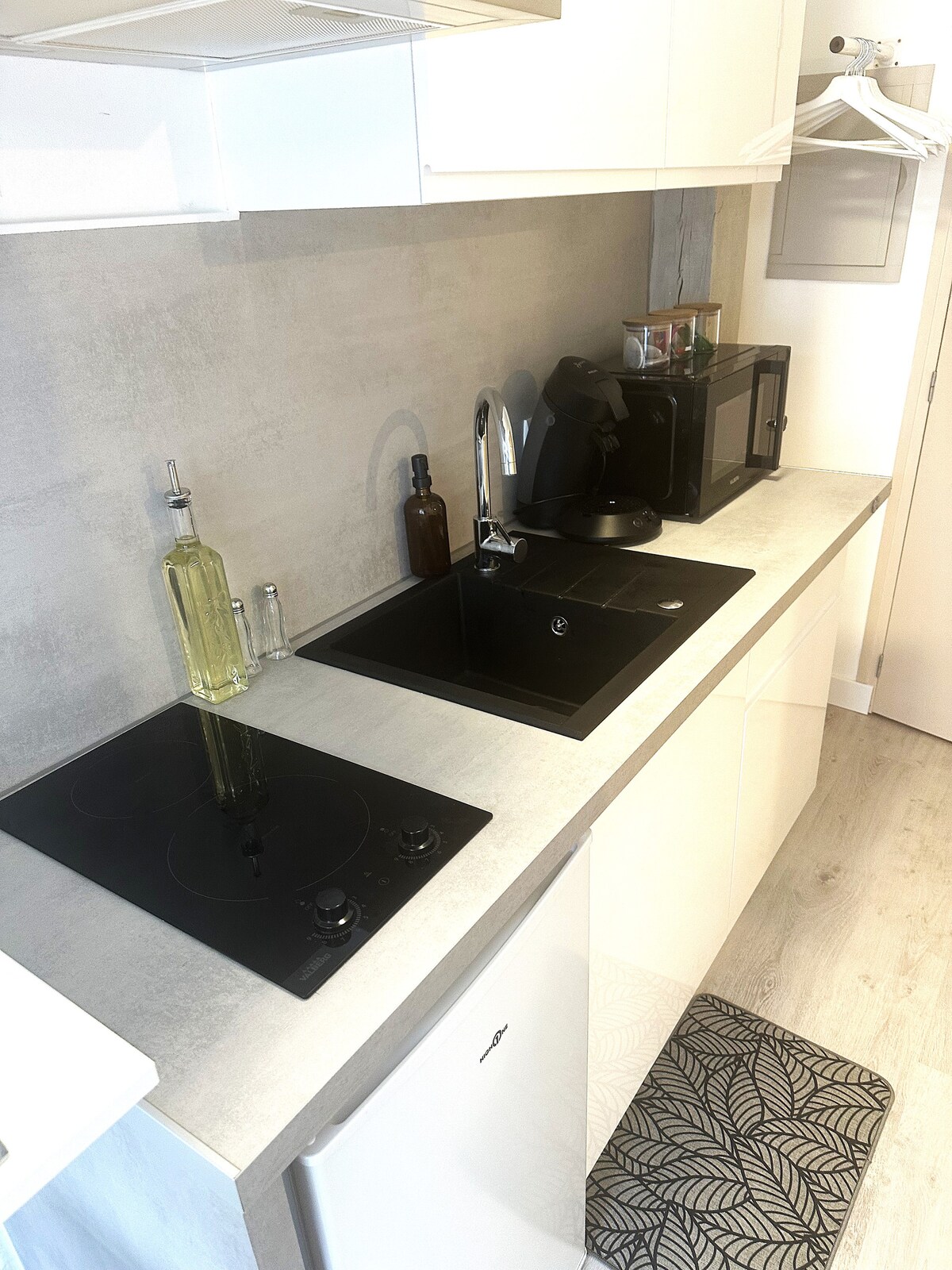 The kitchen area is equipped with a black induction cooktop and a sleek, modern sink. Appliances include a microwave, a coffee machine, and a compact refrigerator. A bottle of olive oil and a small vase subtly adorn the countertop, enhancing the functional yet stylish design.