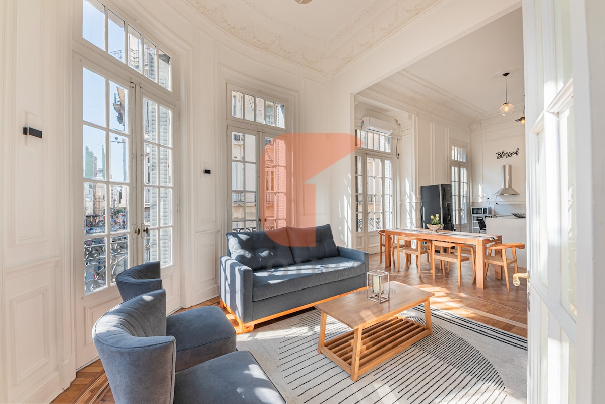 A bright living area features large windows permitting ample natural light. Light wooden flooring complements modern furnishings, including a comfortable sofa and armchairs. A wooden dining table is visible in the background, with a kitchen area showcased to the side. The elegant architecture includes detailed wall moldings and high ceilings.