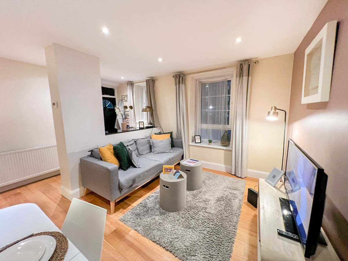 A modern living area is featured, showcasing a comfortable gray sofa adorned with colorful cushions. Light wood flooring enhances the space, while a distinct area rug adds texture. A small coffee table holds various items, and a television is positioned against a pale wall.