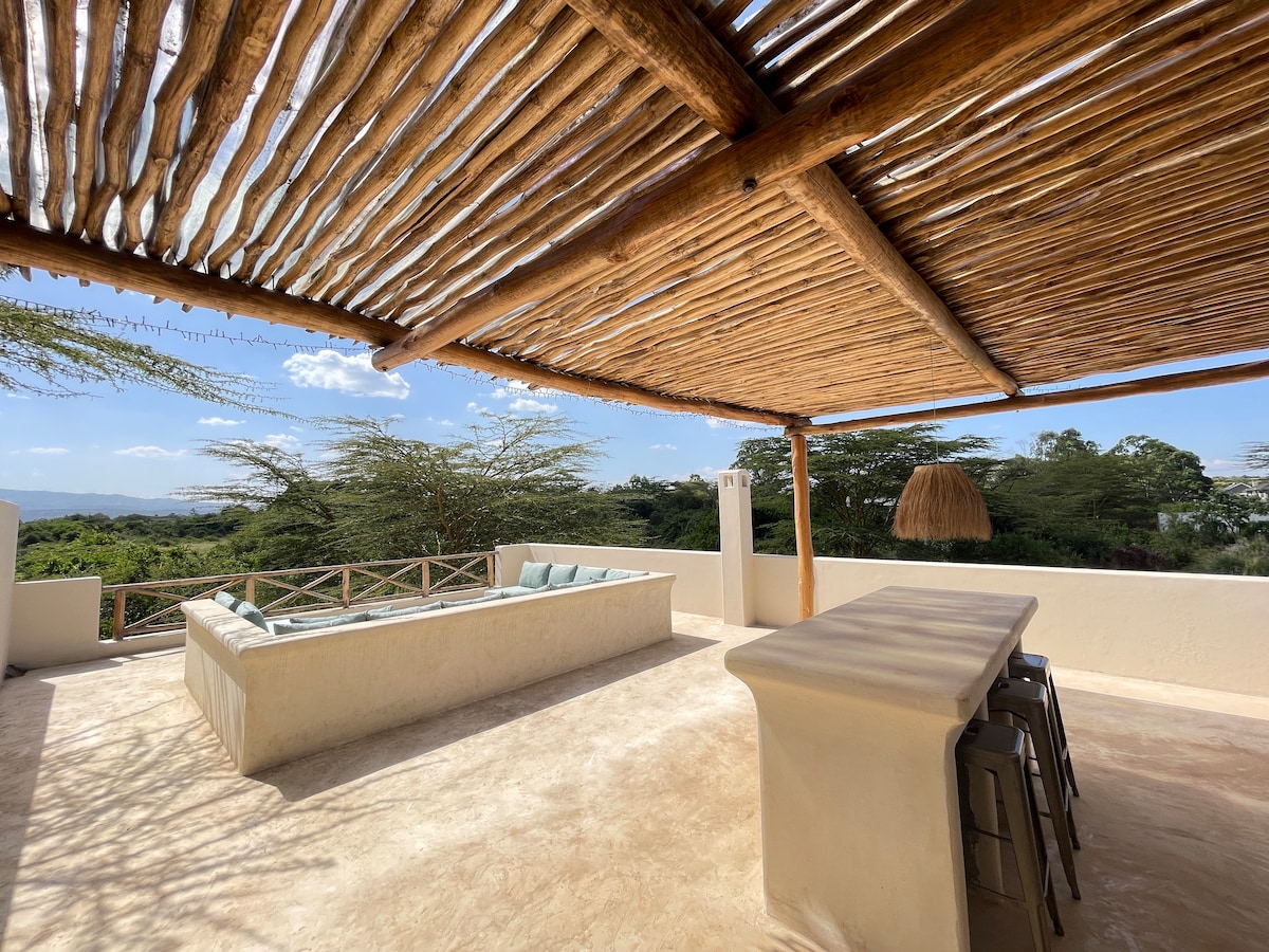 An outdoor terrace is featured, showcasing a spacious lounge area with comfortable seating and a bar counter. The area is shaded by a wooden canopy, while expansive views of the surrounding landscape and greenery are visible beyond.