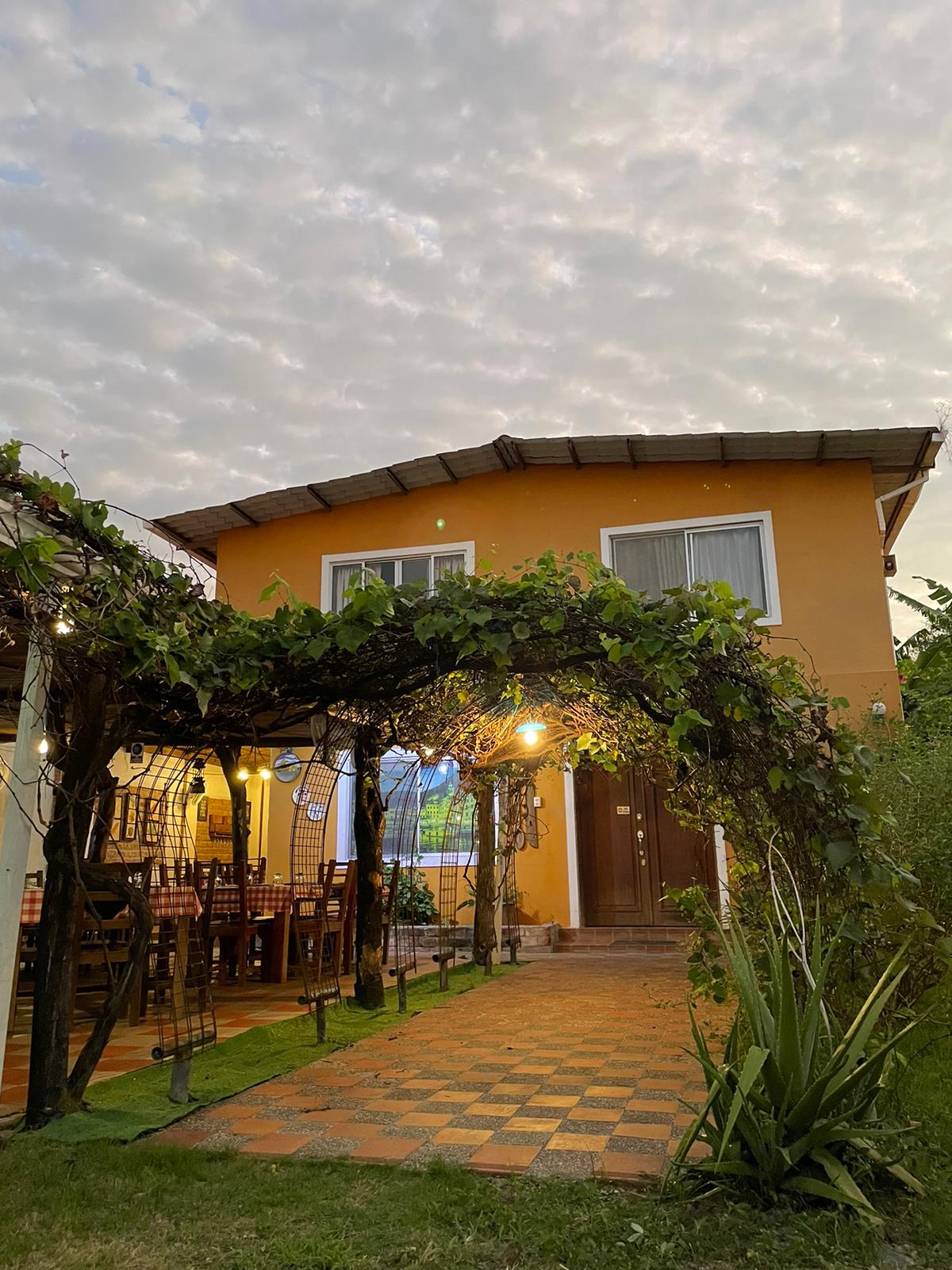 A charming two-story building is framed by a canopy of greenery, with warm light spilling from the entrance. The exterior features earthy tones, and a patterned pathway leads to the doorway, creating an inviting approach surrounded by lush vegetation.
