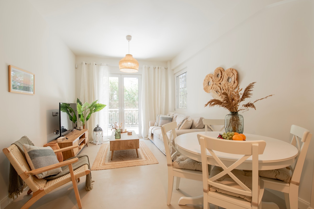 A bright living area is illuminated by natural light through large windows. A comfortable sofa and armchair are arranged around a central coffee table. A dining table with chairs is positioned nearby, complemented by decorative elements including plants and a minimalist light fixture.