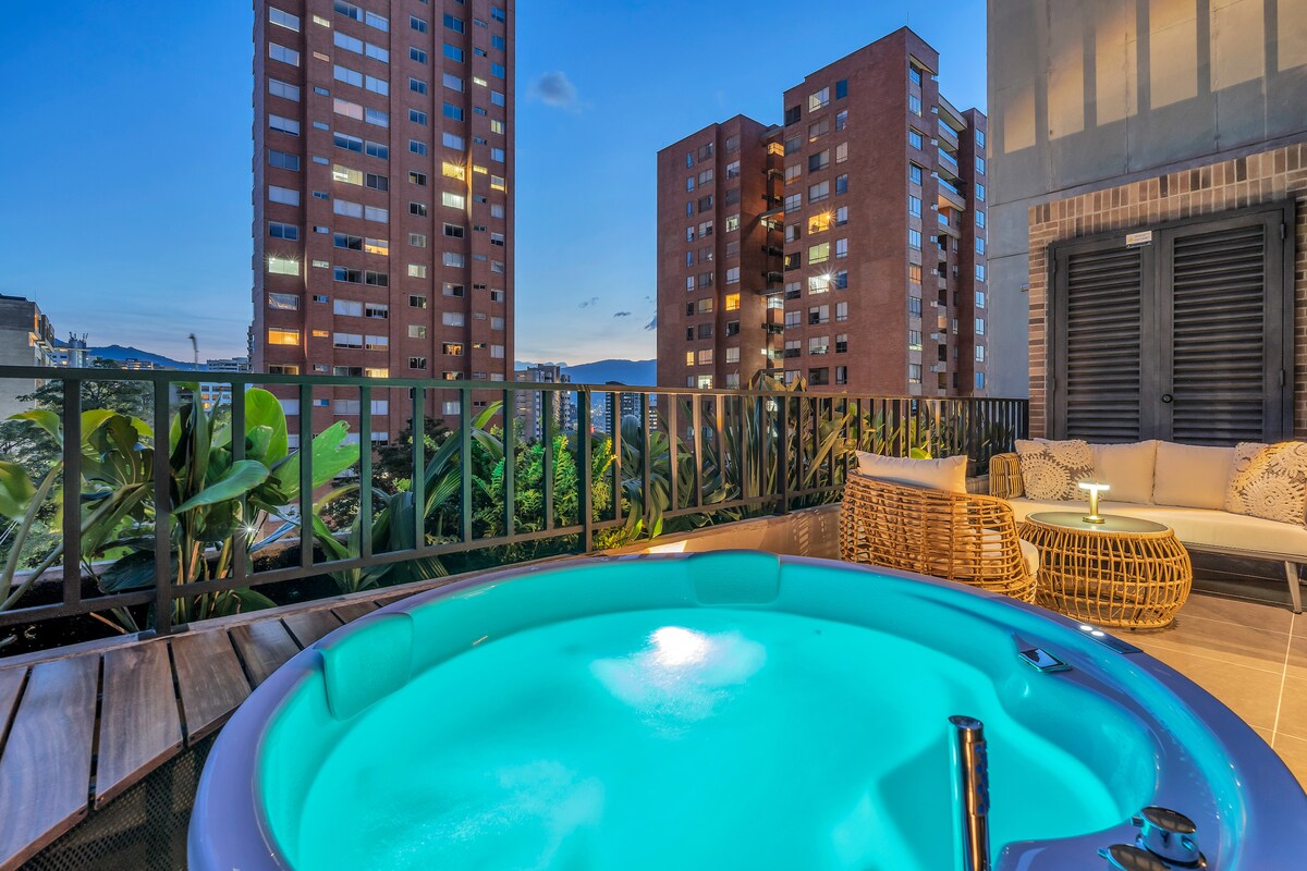 A private jacuzzi offers a calming turquoise glow, surrounded by natural greenery and modern seating on a spacious terrace. Views of surrounding buildings and the evening sky create a serene outdoor setting for relaxation.
