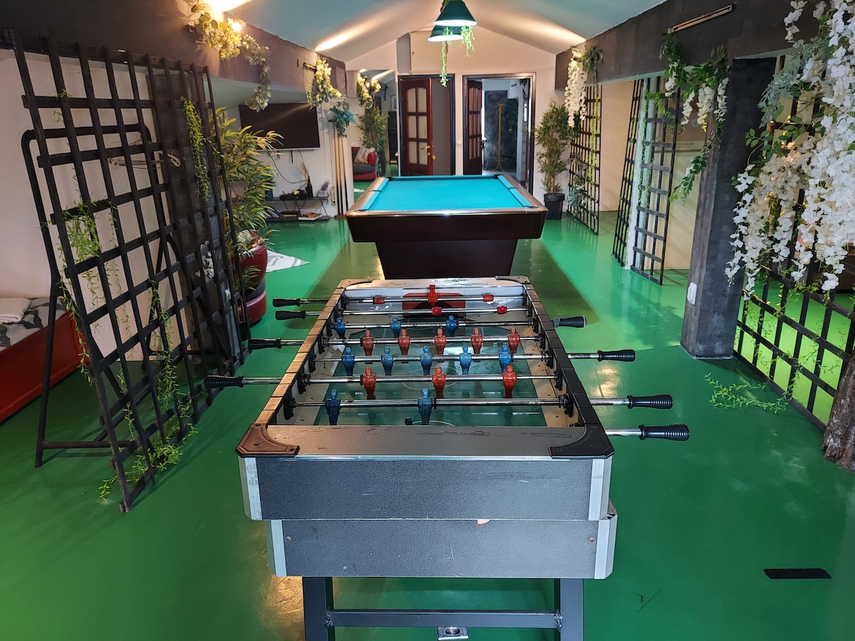 The image displays a spacious recreation area featuring a foosball table in the foreground. In the background, a billiards table is visible under a hanging light. The room is decorated with greenery along the walls, creating a lively atmosphere.