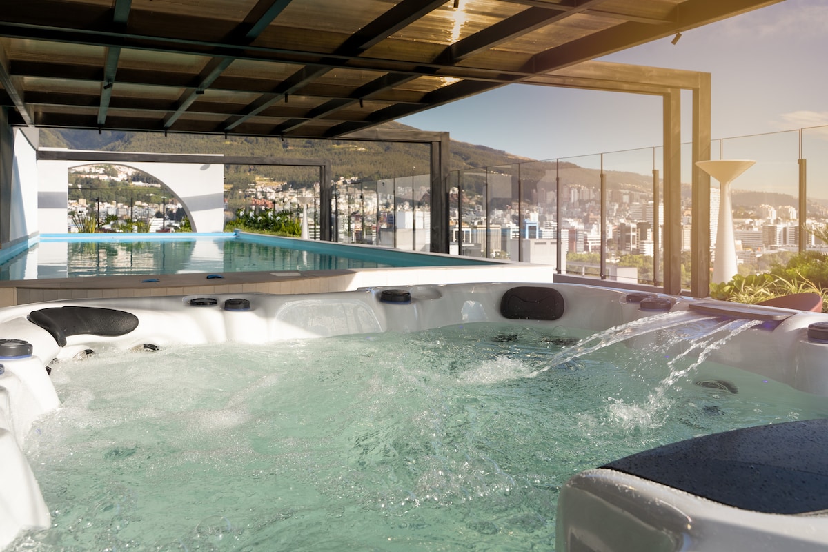 A modern outdoor jacuzzi is surrounded by a sleek deck area, offering a view of the distant mountains and cityscape. The jacuzzi features comfortable seating and bubbling water, inviting relaxation while enjoying the scenic surroundings.