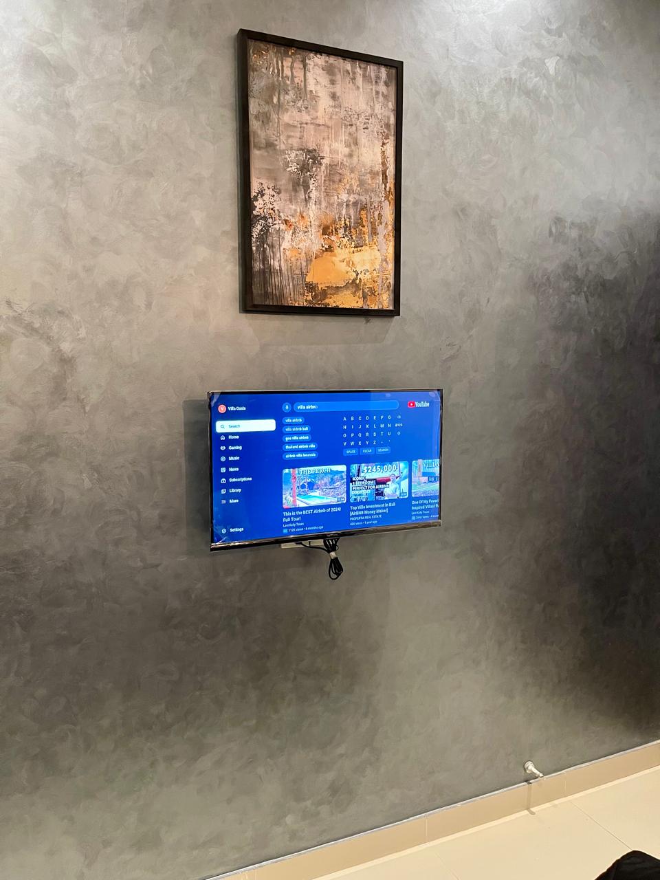 A smart TV is mounted on a textured gray wall, displaying various streaming options. Above the TV, a framed piece of abstract art adds a splash of color. The floor is light-colored, complementing the overall modern aesthetic of the space.