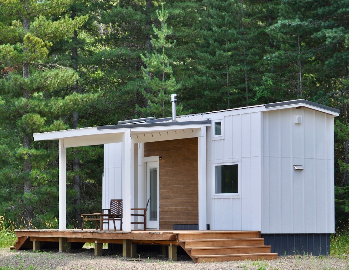 A modern tiny home is surrounded by tall trees, featuring a deck with wooden steps leading to a covered porch. A single chair sits on the porch, providing a designated space for outdoor relaxation.