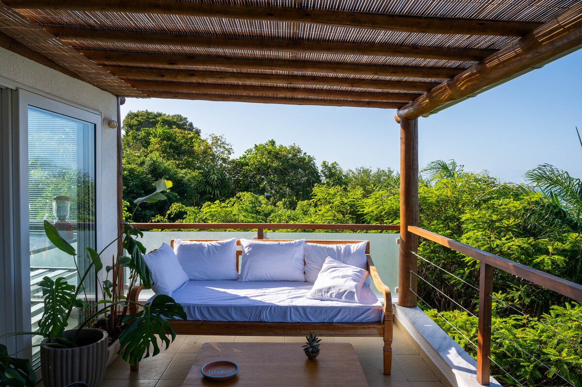 A shaded terrace area features a wooden sofa adorned with white cushions, inviting relaxation. A wooden table is present, alongside lush green plants. The surrounding foliage offers a sense of privacy and a glimpse of distant views, creating a serene outdoor space.
