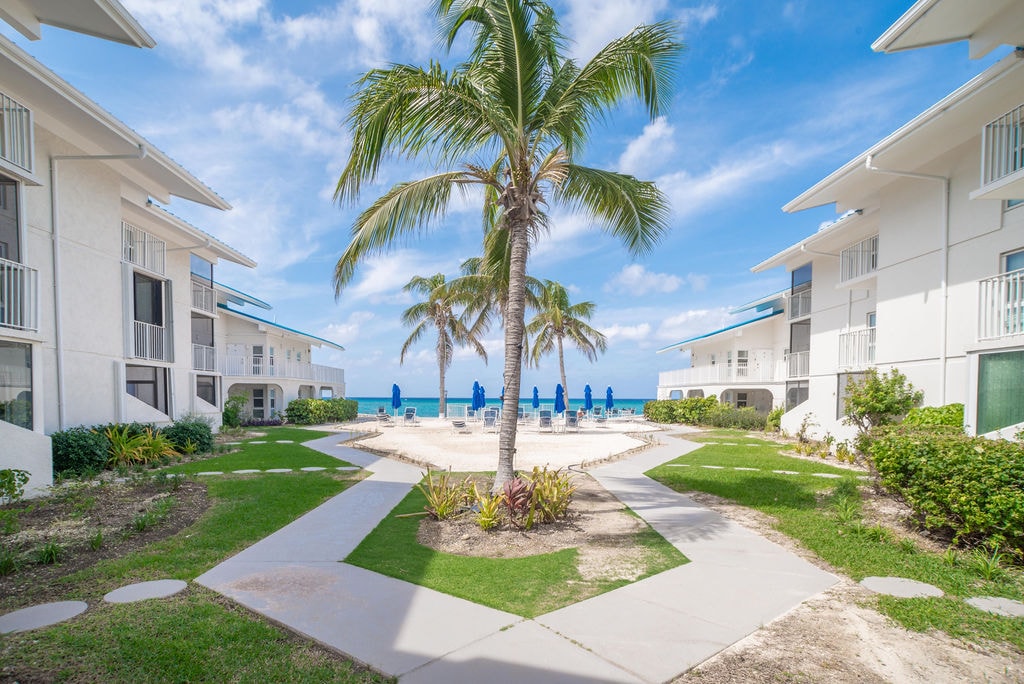 A serene courtyard is adorned with vibrant greenery and swaying palm trees, framing a sandy area. The background features two white buildings with balconies, while blue lounge chairs are positioned along the shoreline, inviting relaxation under the clear blue sky.