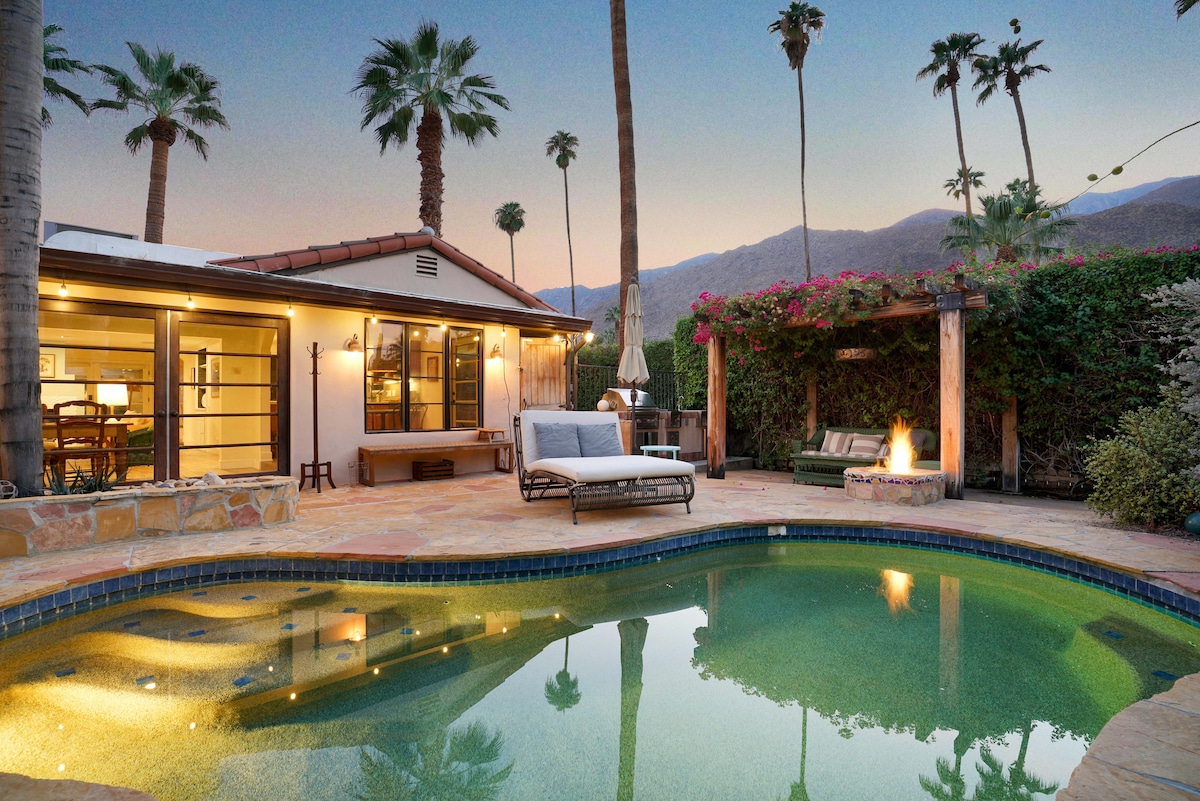 An inviting outdoor space features a heated pool surrounded by lush palm trees and vibrant flora. A cozy fire pit sits nearby, with comfortable seating and soft lighting enhancing the serene atmosphere. The backdrop showcases the mountains under a twilight sky.